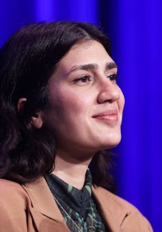 A close-up photograph of a softly smiling brunette woman in a tan blazer, sitting in front of a blue curtain.