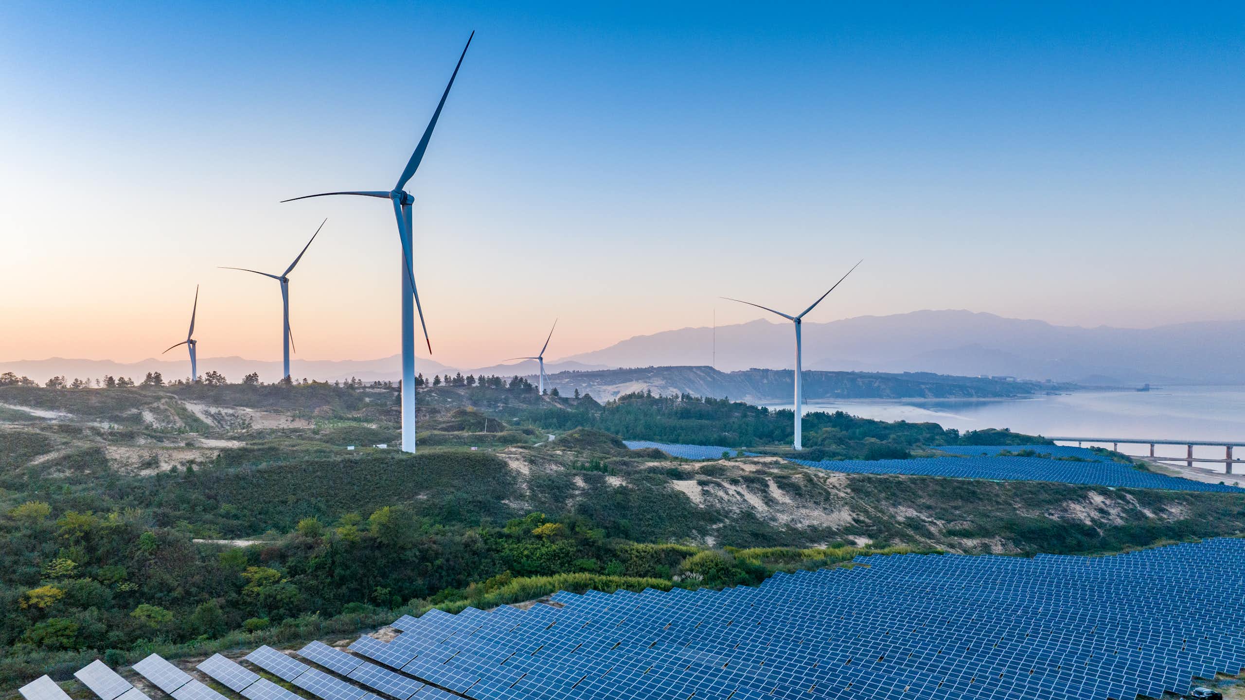 Rolling hills with giant sets of solar panels in lines of about 30 and several big wind turbines dotted over them
