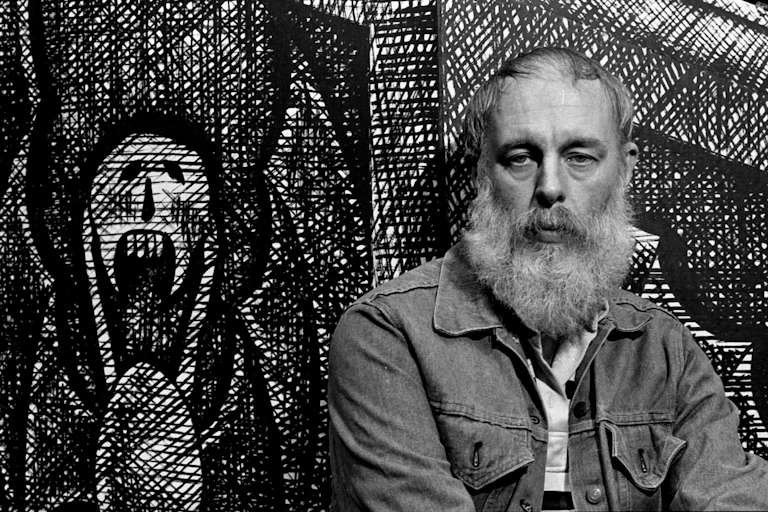 Black and white portrait of bearded, middle-aged man posing next to a drawing of a skeleton-like figure with wings.