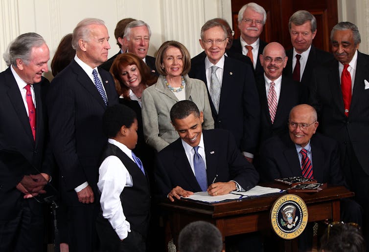 President Barack Obama signs a document, surrounded by members of Congress.