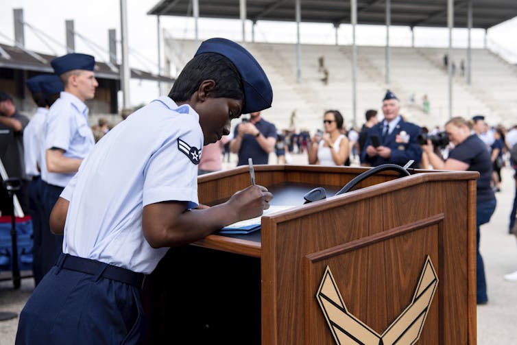 A black woman wearing a blue shirt and blue military cap signs a piece of paper on a lecturn.
