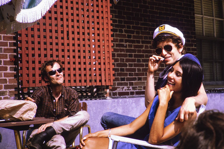 Young man wearing sunglasses smokes cigarette with his arm around a young woman with straight black hair.