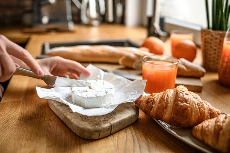 Woman cutting soft cheese on board, with croissants, juice and baguettes on table