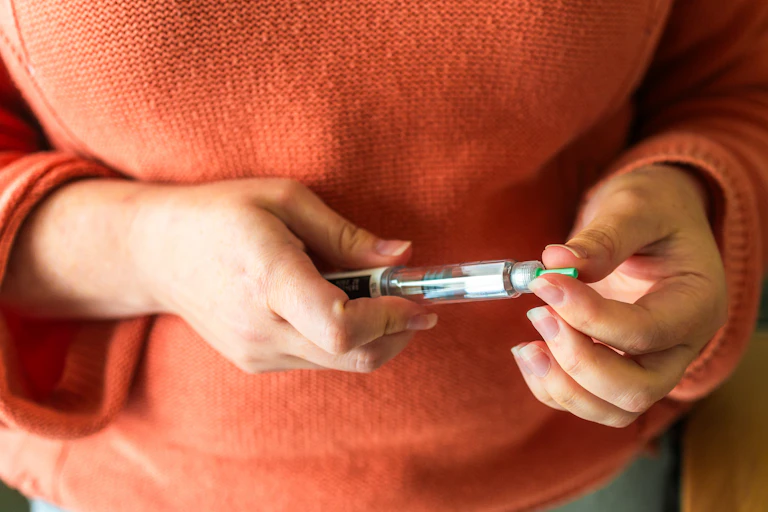 A woman wering an orange jumper holds a weight-loss injection in front of her stomach.
