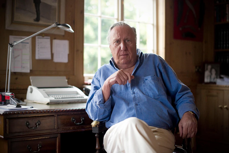 An older well-to-do serious-looking man dressed in chinos and a blue shirt at his desk and typewriter somewhere expensive.