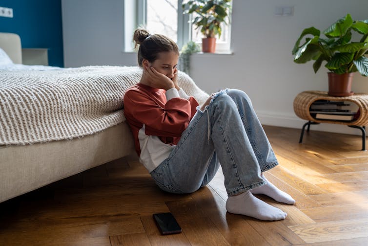 A teenage girl sits on the floor of her bedroom looking sad.
