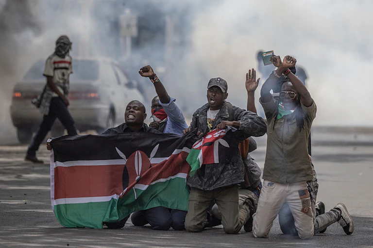 Four men on their knees on a street holding a flag with black, white, red and green stripes with a cloud of smoke behind them