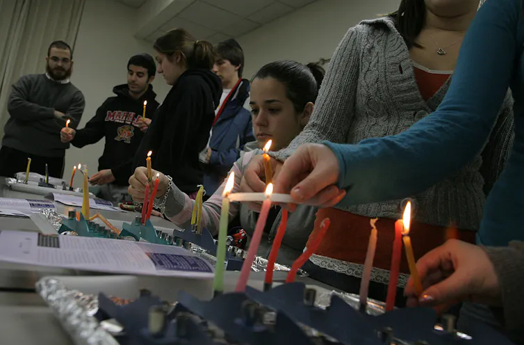 Half a dozen students sit and stand around a table where they are lighting thin colored candles.