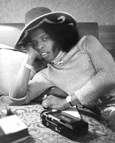 Black man with afro and hat reclining on bed with a tape recorder in front of him.