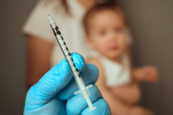 A hand holds a vaccine needle, and an out-of-focus baby sits on a lap in the background