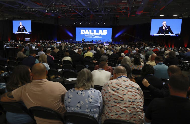 A large audience is seated in a darkened hall, listening to a speaker who appears on two large screens at the front, alongside another screen displaying the words 'Dallas Annual Meeting.'