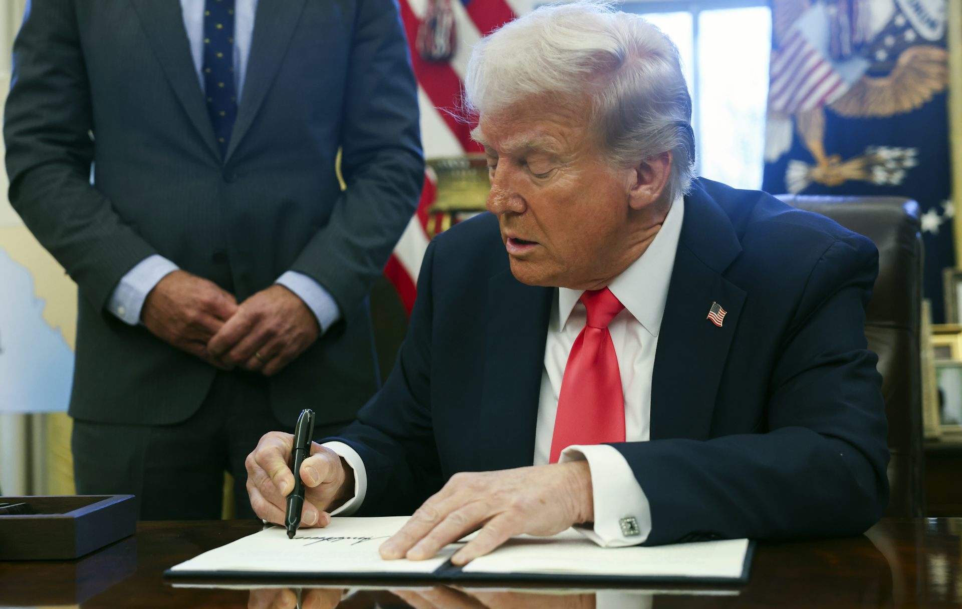An aged white man in a suit and red tie signs a document