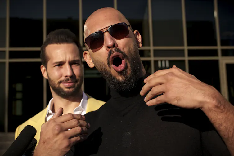 A bald man with a beard and sunglasses gestures toward himself with an open mouth. Another bearded man, this one with hair, stands behind him
