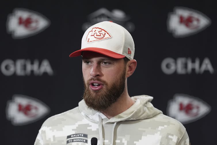A man with a beard in a ballcap speaks into a microphone while sitting down