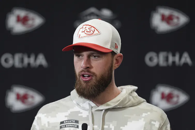 A man with a beard in a ballcap speaks into a microphone while sitting down