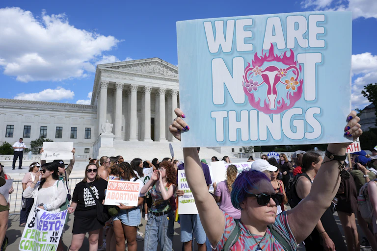 Protesters congregate in front of a white, Hellenistic style buliding. One protester holds a sign that says 'we are not things' with the image of a uterus on it