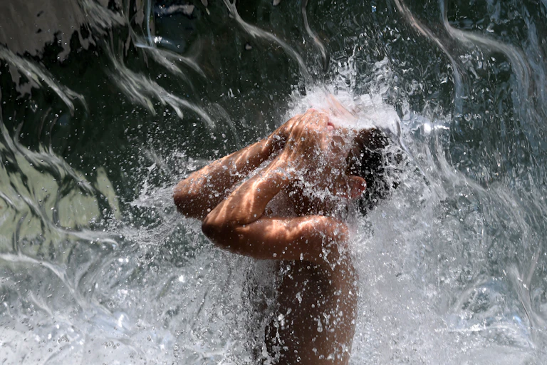 A young man stands under a waterfall and splashes water onto his face. The photo catches the rippling waves of the water falling.