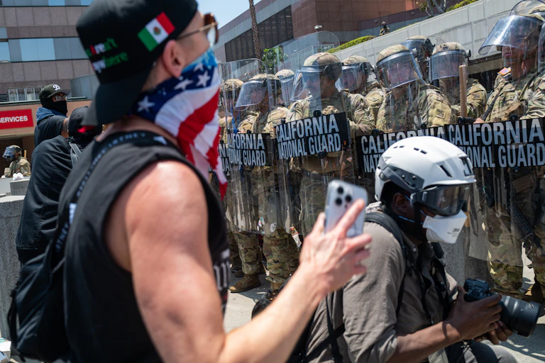Two protesters stand in front of a row of troops wearing helmets and holding body shields.