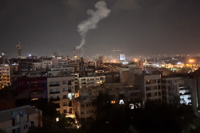 A plume of smoke is seen in a night sky above buildings.