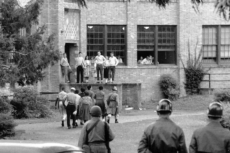 A soldier escorts seven Black students toward a school building as other soldiers watch.