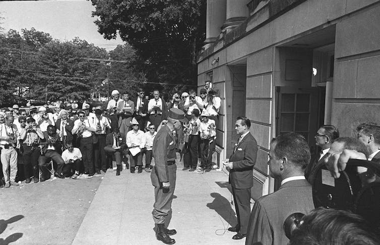 A soldier speaks to a man in a suit in front of a building amid a crowd of onlookers.
