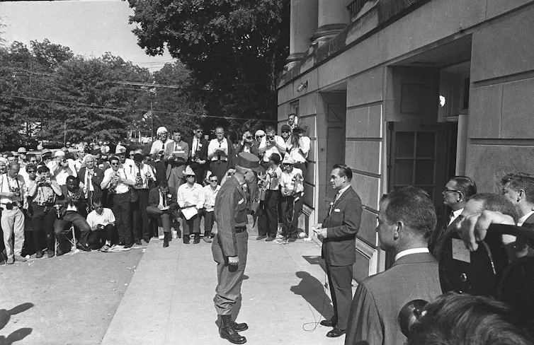 A soldier speaks to a man in a suit in front of a building amid a crowd of onlookers.