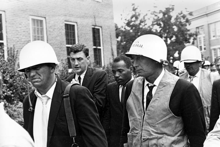 A Black man is escorted toward a building by several men, including three wearing helmets.