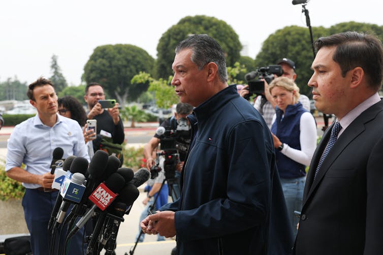 A man speaks in front of a group of microphones, surrounded by other people.
