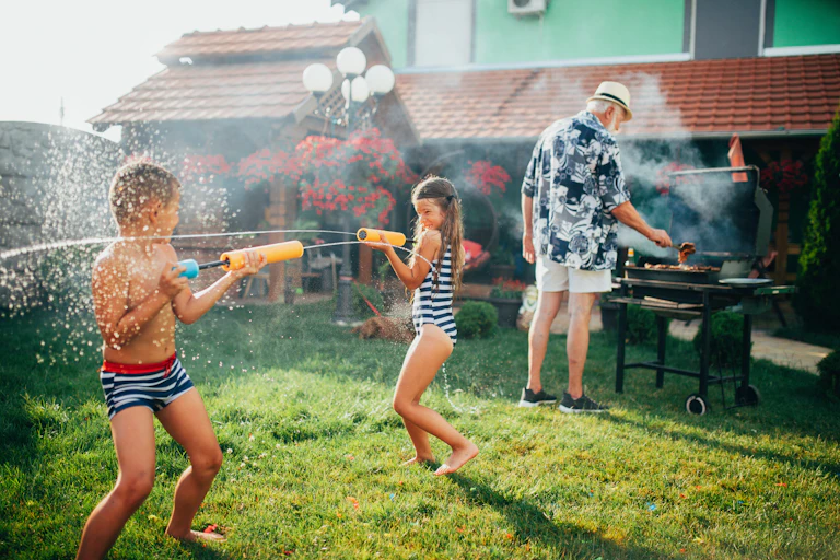 Two children in swimsuits shoot water at one another in the foreground as a grandfather-aged man grills in the background.