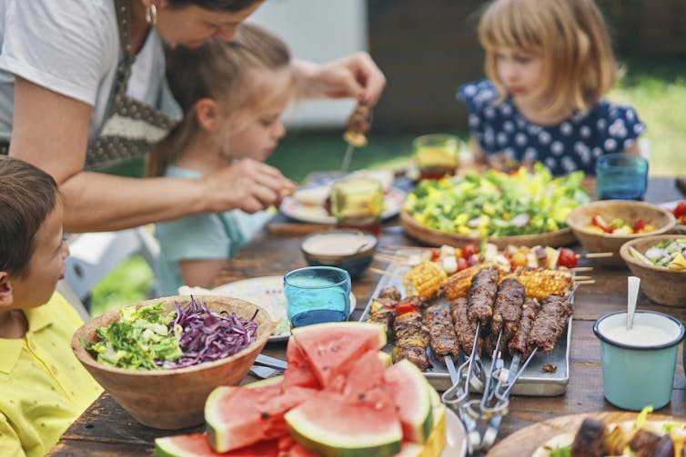 A woman and three young kids sit around an outdoor table eating picnic foods, including watermelon in the foregound.