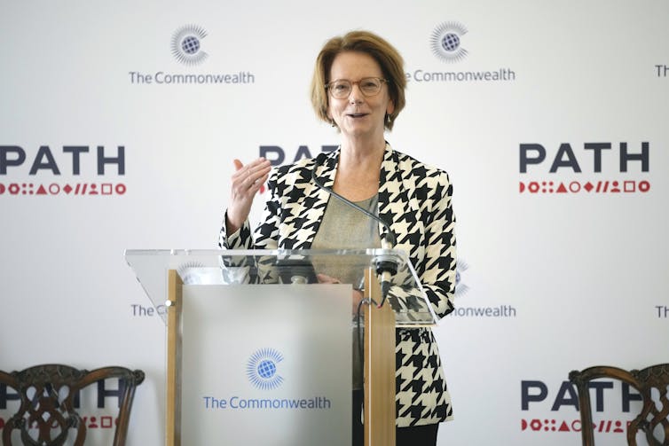 A woman with a short bob in a houndstooth blazer speaks from behind a podium