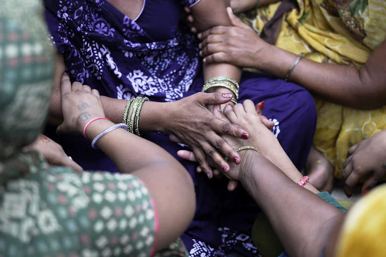A group of hands, arms showing bangles, bodies showing colourful clothing, hold onto each other