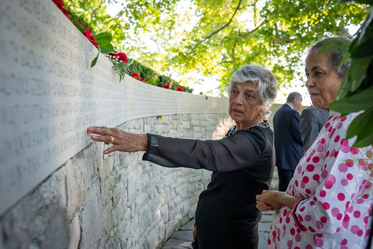 Dos mujeres se paran frente a una pared de concreto bajo, un punto a la inscripción en la pared