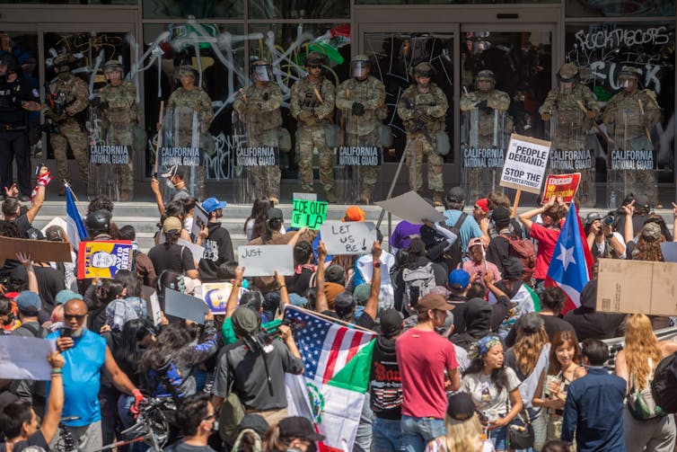 Demonstrators holding signs and flags face members of the military standing guard outside a large building.