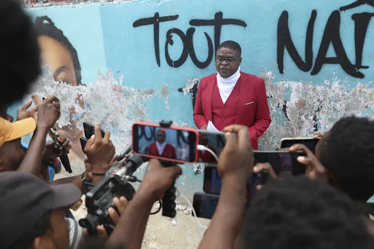 A man in a red suit with glasses stands in front of cameras
