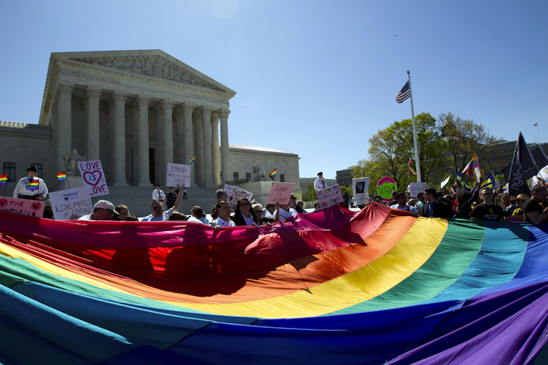 a crowd holds an oversized rainbow flag outside a columned building.