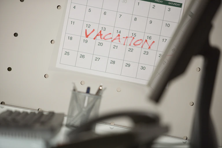 A desk in an office featuring a phone and computer monitor in the foreground, with a calendar hanging on the wall with one week marked off in red text that reads 'Vacation.'