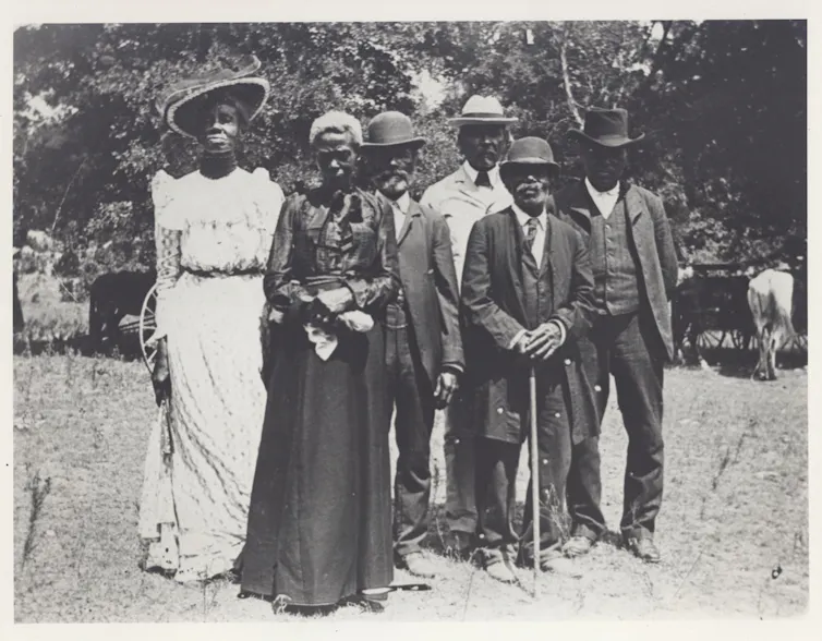 A group of people in fancy dress from the turn of the 19th century, standing together outside.