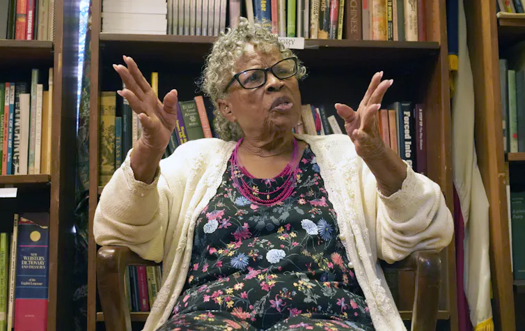 A gray haired woman sitting in a room with bookshelves behind her, gesturing with her hands.