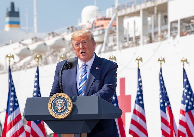 Donald Trump speaking at a podium in front of a row of flags and a naval ship