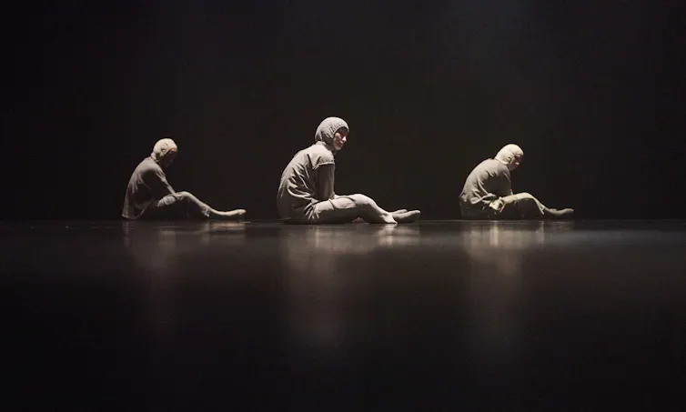 Three people sit on stage in grey tracksuits.