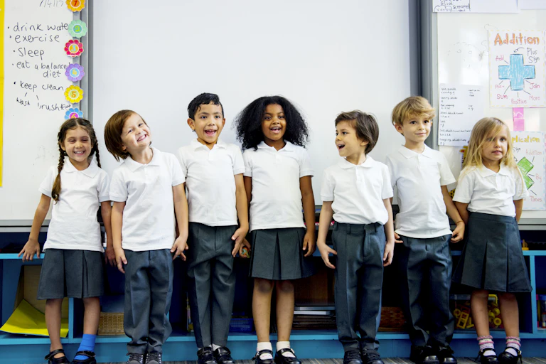A group of young students in uniform line up against a white board in a classroom.