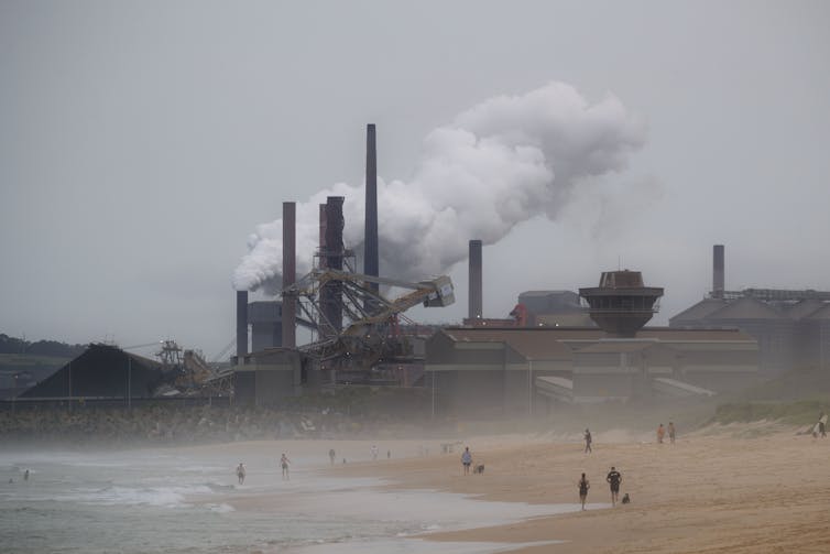 People walk on beach in front of industrial plant billowing steam.