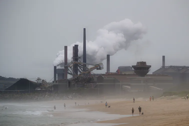 People walk on beach in front of industrial plant billowing steam.