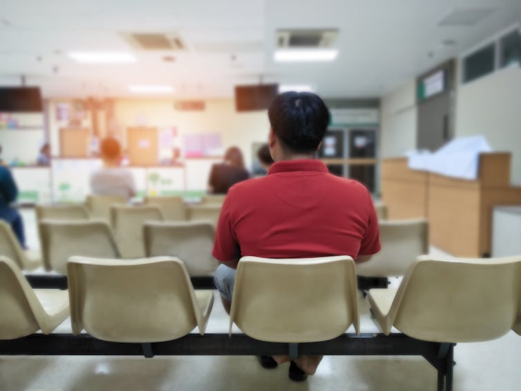 In a red shirt, a man who is sitting in the waiting room at the hospital is seen from behind.