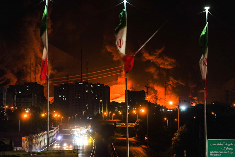 Flames from a mnissile attack on an Iranian oil refinery with Iranian flags in the foreground.