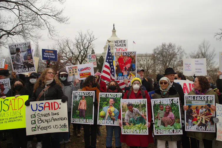 Protesters with signs featuring photos of aid workers that read 'the faces of foreign aid'