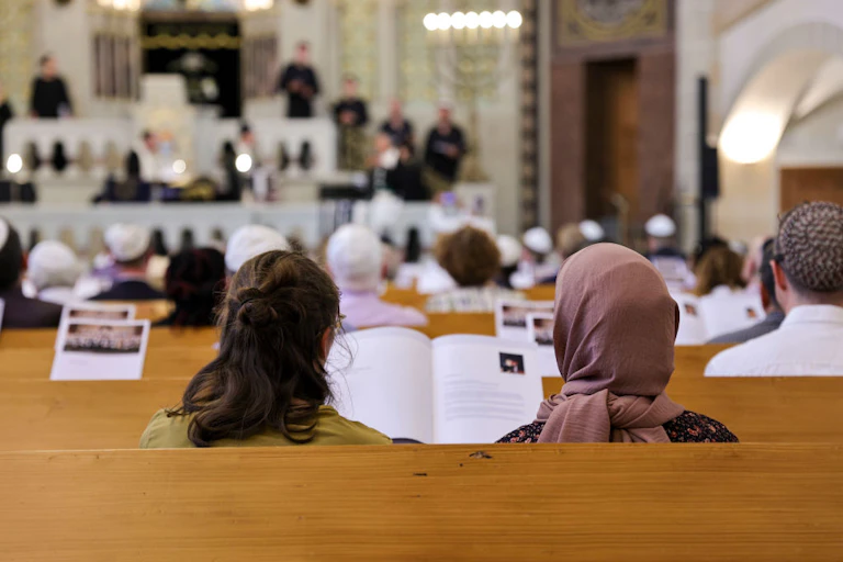 Two women, one of whom is wearing a purple head covering, sit next to each other in a pew, viewed from the back.