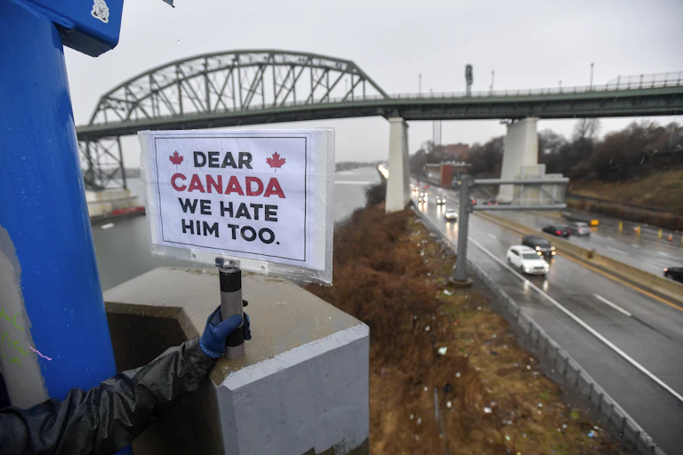 A person holds a sign reading Dear Canada, We Hate Him Too with a bridge in the background on a cloudy day.
