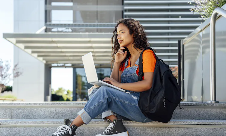 A woman sits on steps while holding a laptop and wearing a backpack.
